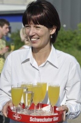 Eine Frau hält einen roten Tablett mit Gläsern voller Orangensaft, lächelt und schaut in die Kamera. Sie trägt ein weißes Hemd. Das Tablett hat den Text 'Schwechater Bier' darauf.