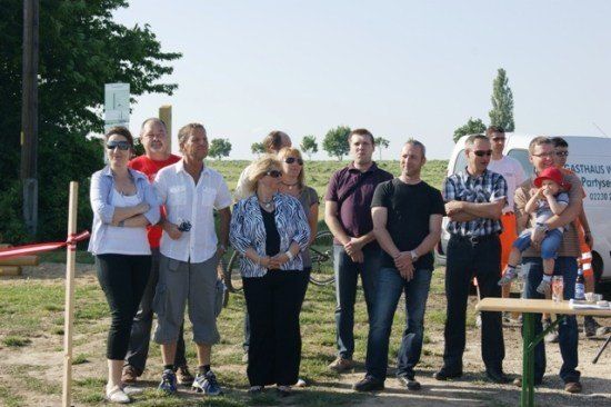Eine Gruppe von Menschen steht auf einem Feld mit einem weißen Van und einem Holztisch im Hintergrund. Sie sind lässig gekleidet und warten scheinbar auf etwas.