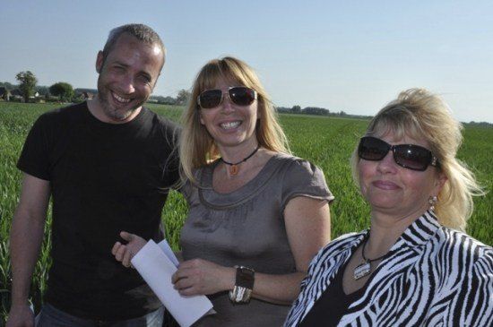 Drei Personen stehen in einem Feld und lächeln für ein Foto. Ein Mann in Schwarz, eine Frau in Grau mit einem Papier und eine Frau in einem Zebra-Streifen-Jacke.