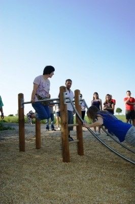 Zwei Frauen trainieren an einem hölzernen Klimmzug-Stab in einem Outdoor-Spielplatz, während mehrere Menschen im Hintergrund zusehen.