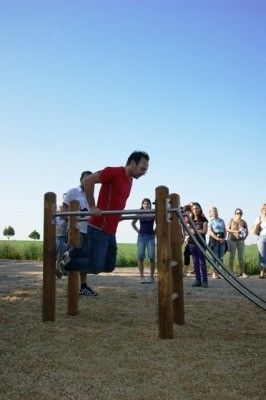Ein Mann in einem roten T-Shirt und blauen Jeans macht Klimmzüge an einer Holzstange im Freien, während mehrere Leute hinter ihm stehen und zusehen.
