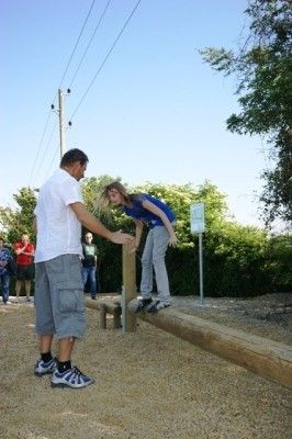 Ein Mann hilft einer Frau, auf einem schmalen Holzbalken in einem Outdoor-Spielplatz zu balancieren.
