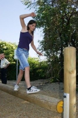 Eine Frau balanciert auf einem Holzbalken im Freien, trägt ein lila Tanktop, Jeansshorts und weiße Turnschuhe. Hinter ihr steht ein Mann in der Nähe eines Schildes und Bäumen.