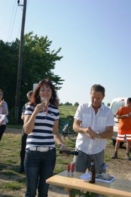 Eine Frau in einem gestreiften Shirt hält ein Weinglas und steht neben einem Mann in einem weißen Shirt, beide blicken auf einen Tisch mit Flaschen und Essen, mit einem Fahrrad und einem weißen Van im Hintergrund.