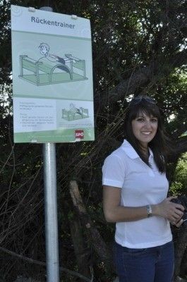 Eine Frau in einem weißen Shirt und Jeans steht neben einem Pfosten mit einem Schild, das Ruckentrainer besagt. Sie lächelt und hält eine Kamera.