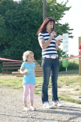 Eine Frau mit einem Mikrofon steht neben einem kleinen Mädchen im Park, mit Bäumen und einer Bank im Hintergrund.