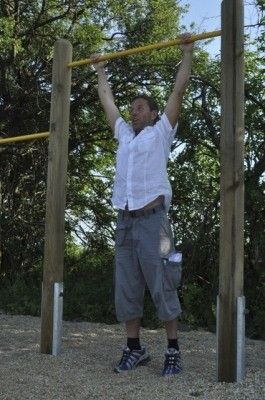 Ein Mann in einem weißen Hemd und grauen Shorts führt Klimmzüge an einer Holzstange in einer Außenumgebung mit Bäumen im Hintergrund durch.