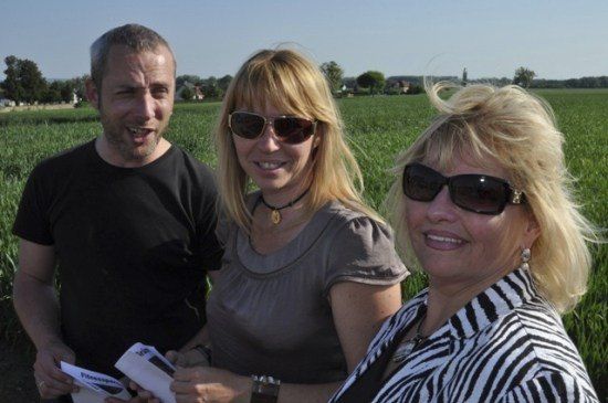 Drei Menschen stehen auf einem Feld, lächeln und halten Papiere in den Händen. Der Mann auf der linken Seite trägt ein schwarzes T-Shirt, die Frau in der Mitte trägt ein graues Oberteil und die Frau auf der rechten Seite trägt ein Zebra-gestreiftes Hemd. Sie tragen alle eine Sonnenbrille.