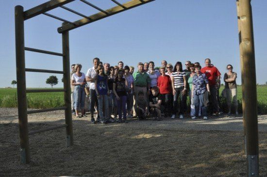 Eine Gruppe von Menschen steht vor einer Holzstruktur in einem Außenbereich. Einige tragen Brillen und einige tragen Turnschuhe. Sie posieren wahrscheinlich für ein Foto. Hinter ihnen ist ein Feld mit Gras und Bäumen. Der Himmel ist blau und klar.
