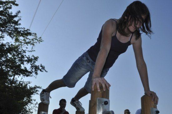 Eine junge Frau macht einen Klimmzug an einer horizontalen Stange im Freien. Sie trägt ein schwarzes Tanktop und Jeansshorts. Im Hintergrund stehen zwei Personen in der Nähe eines Baumes.