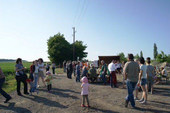 Eine Gruppe von Menschen, einschließlich Erwachsener und Kinder, versammelt sich in einem Außenbereich, möglicherweise für eine Veranstaltung, mit einem Schuppen und Bäumen im Hintergrund.