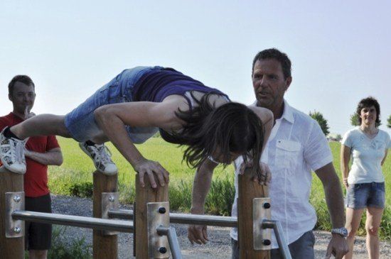 Eine Frau führt einen Handstand auf einer horizontalen Stange durch, während zwei Personen von hinten zusehen. Der Ort ist draußen, mit einem Grasfeld und einem klaren Himmel im Hintergrund.