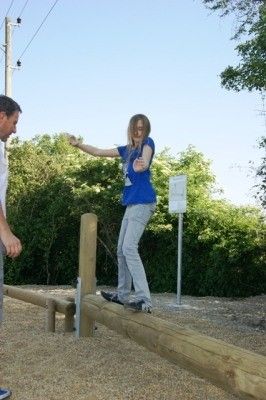 Eine Frau in einem blauen Shirt und grauer Hose balanciert auf einem Holzbalken im Freien, während ein Mann zusieht. Bäume und ein Versorgungsmast sind im Hintergrund.