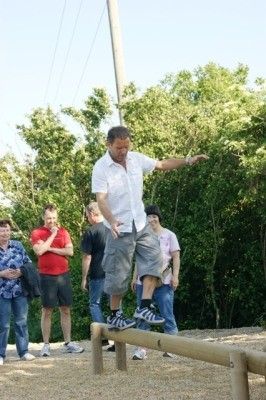 Ein Mann in einem weißen Hemd und grauen Shorts balanciert auf einer horizontalen Stange, während andere in einer Außenumgebung zusehen.