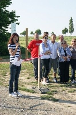 Eine Gruppe von Menschen steht bei einer Schleifenschneidezeremonie mit einer Frau, die ein Mikrofon und ein Stück Papier in der Hand hält. Sie befinden sich auf einem Feld aus Erde mit Bäumen im Hintergrund.