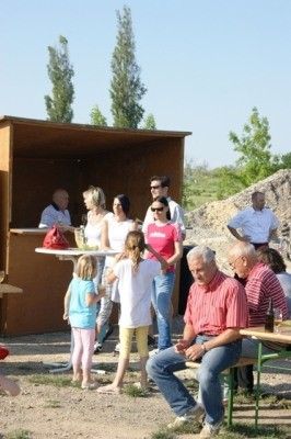 Eine Gruppe von Menschen, einschließlich Kindern, versammelt sich um einen kleinen Holzstand mit einem Tisch und einer roten Tasche darauf. Einige sitzen, während andere stehen, und es gibt Bäume und einen Haufen Erde im Hintergrund.