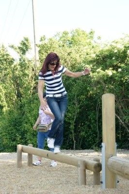 Eine Frau in einem gestreiften Shirt und Jeans springt über eine Holz-Hürde, während ein Kind hinter ihr steht.
