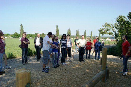 Eine Gruppe von Menschen steht in einem Schotterbereich, wahrscheinlich auf einer Tour, mit einem Feld und Bäumen im Hintergrund.