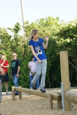 Ein Mädchen in einem blauen Shirt balanciert auf einem Holzbalken in einem Park, mit mehreren Leuten, die hinter ihr stehen, und Bäumen im Hintergrund.