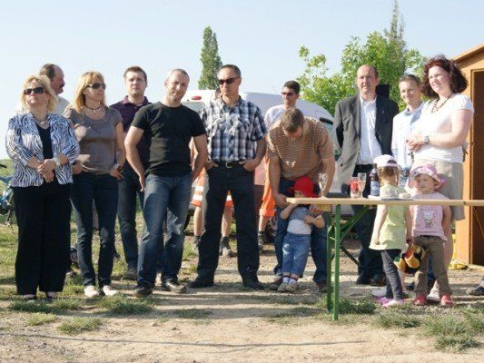 Eine Gruppe von Menschen, einschließlich Erwachsener und Kinder, steht in einem Außenbereich. Einige tragen Sonnenbrillen, und ein Kind beugt sich über einen Tisch.