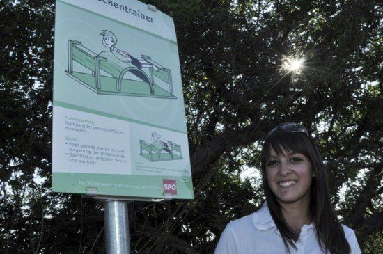 Eine Frau steht neben einem Schild mit einer Zeichnung einer Person auf einem Trampolin und lächelt in die Kamera. Das Schild hat Text auf Deutsch und ein Logo.