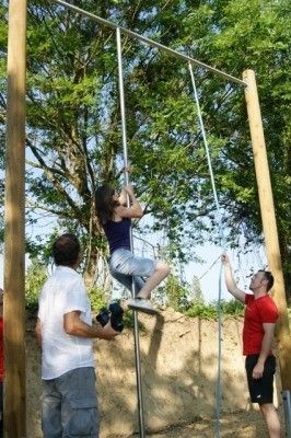 Eine Frau klettert eine Seilleiter hinauf, während zwei Männer das Seil unter ihr halten. Sie befinden sich in einer Außenumgebung mit Bäumen und Erdboden.
