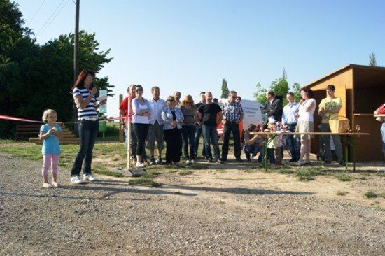 Eine Gruppe von Menschen steht im Freien in einer Reihe und hört einer Frau zu, die in ein Mikrofon spricht. Ein Tisch mit einem Korb steht in der Nähe.