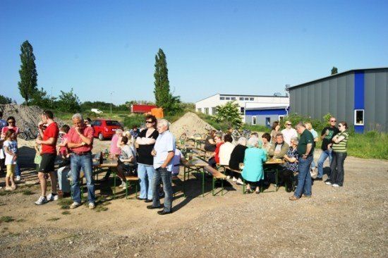 Eine Gruppe von Menschen versammelt sich um Tische in einem Außenbereich, möglicherweise für eine Gemeinschaftsveranstaltung. Einige sitzen, während andere in der Nähe stehen. Im Hintergrund sind Gebäude und ein rotes Auto zu sehen.