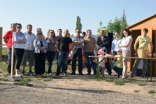Eine Gruppe von Menschen steht draußen, einige in der Nähe eines Tisches mit einem Korb und einem Weinglas, andere in der Nähe eines Bandes und eines kleinen Gebäudes. Bäume und ein Fahrzeug sind im Hintergrund.