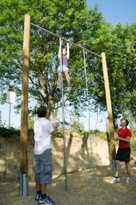 Drei Personen sind an einem sonnigen Tag draußen. Eine Person klettert an einer vertikalen Stange, während zwei andere zusehen. Der Bereich ist von Bäumen umgeben und hat einen sandigen Boden.