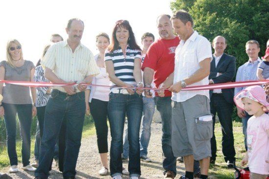Eine Gruppe von Menschen versammelt sich im Freien für eine Bandzeremonie. Zwei Männer und zwei Frauen halten ein rotes Band, während andere zusehen.