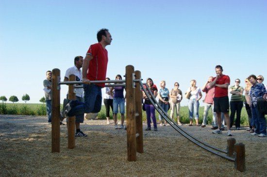 Ein Mann in einem roten T-Shirt führt Klimmzüge an einem Barren in einem Outdoor-Spielplatz durch, während eine Gruppe von Menschen zusieht.