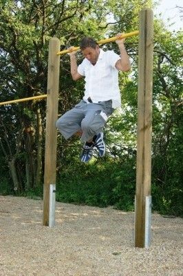 Ein Mann in einem weißen Hemd und grauen Shorts führt Klimmzüge an einer Holzstange in einer Außenumgebung mit Kiesboden und Bäumen im Hintergrund durch.