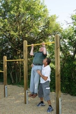 Ein Mann macht Klimmzüge an einer Holzstange, während ein anderer Mann ihn von unten stützt, in einem Außenbereich mit Kiesboden und Bäumen im Hintergrund.