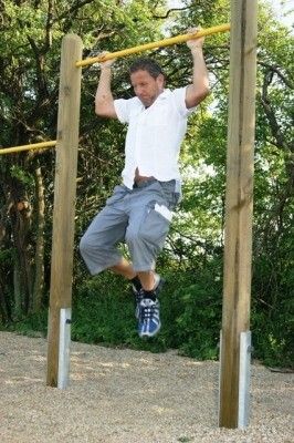Ein Mann in einem weißen Hemd und grauen Shorts macht Klimmzüge an einer Holzstange in einem Außenbereich mit Kiesboden und Bäumen im Hintergrund.