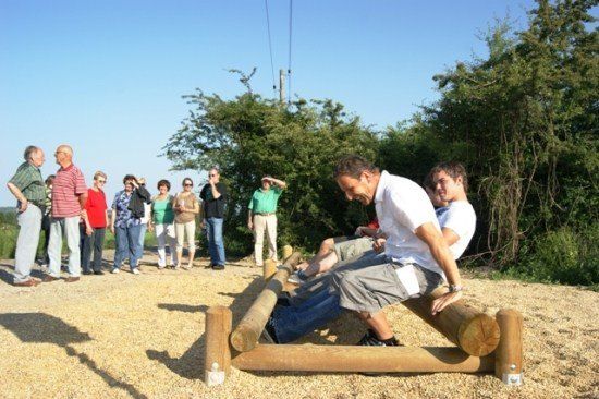 Eine Gruppe von Menschen beobachtet zwei Männer auf einem hölzernen Hindernisparcours. Die Männer sitzen auf Baumstämmen und einer lehnt sich nach vorne.