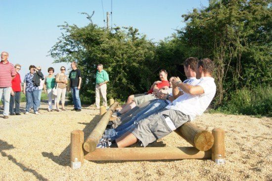 Eine Gruppe von Menschen versammelt sich um einen hölzernen Hindernisparcours auf einer Kiesfläche, wobei einige auf Baumstämmen sitzen und andere dahinter stehen.