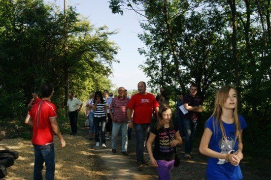 Eine Gruppe von Menschen, darunter Erwachsene und Kinder, geht auf einem mit Bäumen und Grün umgebenen Erdweg. Einige tragen Freizeitkleidung, während andere formellere Kleidung tragen. Einige Personen halten Papiere in den Händen, möglicherweise für eine geführte Tour.