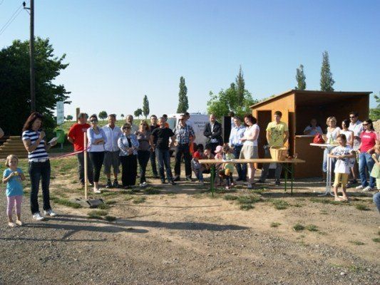Eine Gruppe von Menschen, einschließlich Erwachsener und Kindern, versammelt sich im Freien auf einem Feld. Eine Frau steht in der Nähe eines Bandes, und ein kleines Holzhäuschen ist im Hintergrund zu sehen.