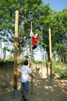 Ein Mann in einem roten T-Shirt und schwarzen Shorts klettert an einer vertikalen Stange in einer Außenumgebung mit sandigem Boden und Bäumen im Hintergrund. Ein anderer Mann in einem weißen T-Shirt und grauen Shorts steht in der Nähe, möglicherweise als Unterstützung.