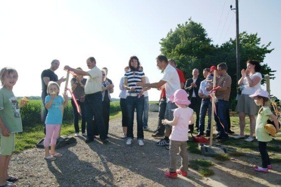 Eine Gruppe von Menschen, einschließlich Erwachsener und Kindern, steht auf einem Kiesweg. Zwei Erwachsene halten rote Bänder, möglicherweise für eine Band-Schneidzeremonie. Die Umgebung ist draußen mit Bäumen und Gras im Hintergrund.