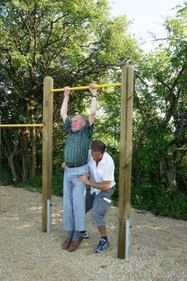 Ein älterer Mann führt Klimmzüge an einer Holzstange in einer Außenumgebung durch, wobei ein anderer Mann ihm hilft. Die Umgebung besteht aus Kies und Bäumen im Hintergrund.