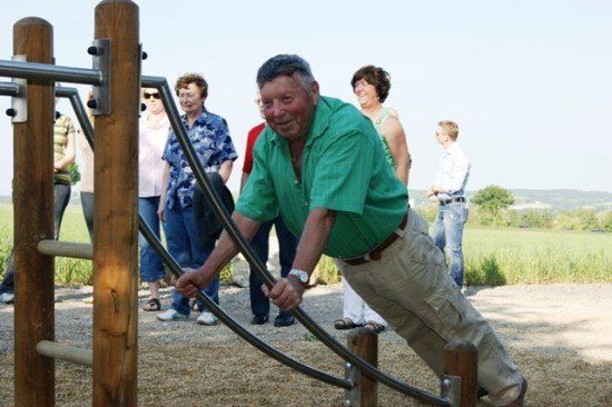 Ein älterer Mann macht Klimmzüge an einem Holz- und Metallapparat, während mehrere Leute zusehen.