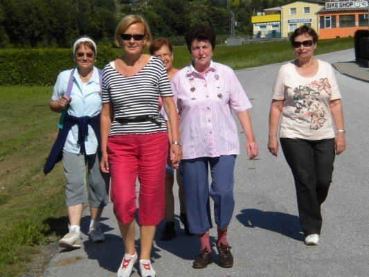 Fünf Frauen gehen auf einem asphaltierten Weg, gekleidet in Freizeitkleidung, mit einem Grasbereich und einem Gebäude im Hintergrund.