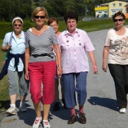 Fünf Frauen gehen auf einem asphaltierten Weg, gekleidet in Freizeitkleidung, mit einem Grasbereich und einem Gebäude im Hintergrund.
