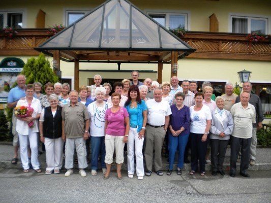 Eine Gruppe älterer Menschen, lässig gekleidet, posiert für ein Gruppenfoto vor einem Gebäude mit einem Glaspavillon.