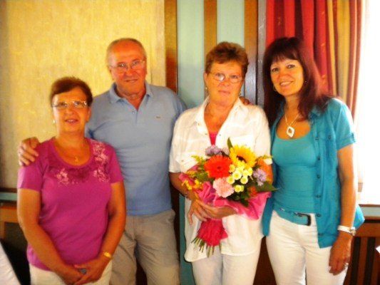 Vier Menschen stehen zusammen in einem Raum. Eine Frau in der Mitte hält einen Blumenstrauß. Der Mann auf der linken Seite lächelt und hat seinen Arm um die Frau auf der linken Seite gelegt. Die Frau auf der rechten Seite lächelt und hat ihre Hand auf der Schulter der Frau in der Mitte.