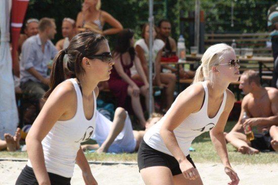 Zwei Frauen in weißen Tank Tops und schwarzen Shorts spielen Beachvolleyball auf einem Sandplatz, mit Zuschauern im Hintergrund.
