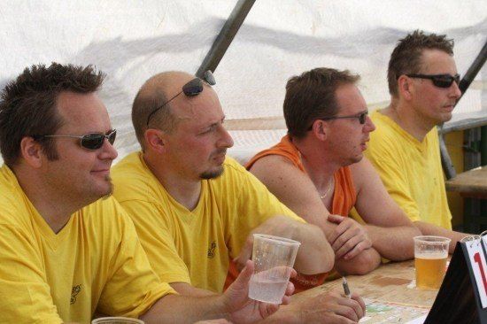 Vier Männer in gelben T-Shirts sitzen an einem Tisch, zwei mit Brillen und zwei mit Getränken, einer schreibt.