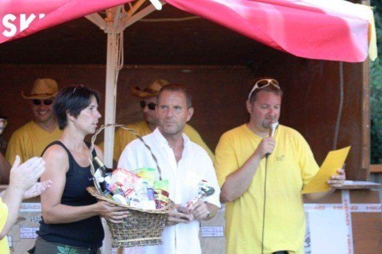 Eine Gruppe von Menschen in gelben T-Shirts, ein Mann hält einen Korb mit Preisen, ein anderer Mann hält ein Mikrofon und eine Frau in einem schwarzen Tanktop.